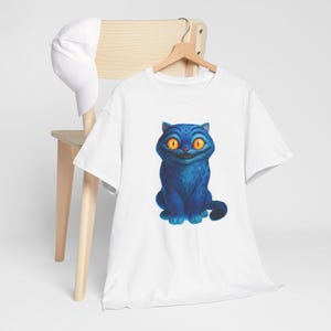 May include: White t-shirt featuring a cartoon blue cat with orange eyes and a wide grin. The cat is sitting upright. The t-shirt is hanging on a wooden hanger. A white baseball cap rests on a wooden chair.