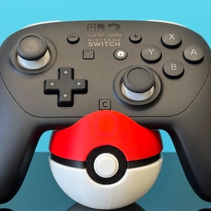 K&ouml;nnte beinhalten: Ein schwarzer Nintendo Switch Pro Controller mit wei&szlig;en Akzenten, der auf einem rot-wei&szlig;-schwarzen Pok&eacute;ball liegt. Der Controller hat X-, Y-, A- und B-Tasten sowie ein Steuerkreuz.