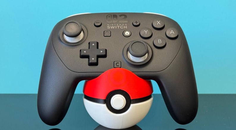 K&ouml;nnte beinhalten: Ein schwarzer Nintendo Switch Pro Controller mit wei&szlig;en Akzenten, der auf einem rot-wei&szlig;-schwarzen Pok&eacute;ball liegt. Der Controller hat X-, Y-, A- und B-Tasten sowie ein Steuerkreuz.
