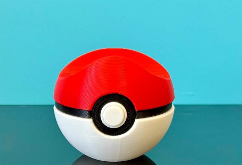 K&ouml;nnte beinhalten: Ein rot-wei&szlig;er Pok&eacute;ball, ein kugelf&ouml;rmiges Ger&auml;t aus dem Pok&eacute;mon-Franchise. Die obere H&auml;lfte ist rot, die untere H&auml;lfte ist wei&szlig;, mit einem schwarzen Band und einem wei&szlig;en Knopf in der Mitte. Der Hintergrund ist t&uuml;rkis.