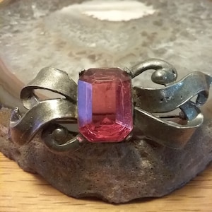 May include: A silver brooch with a pink glass stone in the center. The brooch is shaped like a bow with intricate details.
