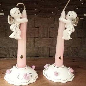 May include: A pair of white ceramic candle holders with pink candles. Each holder features a small angel figurine holding onto the candle. The holders have a scalloped edge and are decorated with pink roses.