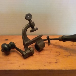 May include: Antique metal tool with a wooden handle, a clamp, and a rotating wheel. The tool is used for leather working.