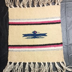 May include: Handwoven beige wall hanging with a geometric design. Features horizontal stripes in pink, black, and gray, and a blue bird-like figure. The bottom and top edges have long, cream-colored fringe.