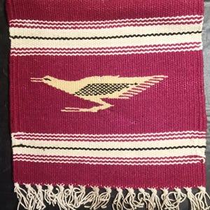 May include: A woven textile in shades of burgundy, cream, and black, featuring a roadrunner design. The rectangular piece has horizontal stripes and fringe along the top and bottom edges. The roadrunner is depicted in cream and black.