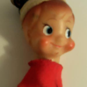 May include: A vintage-style elf ornament featuring a plastic head with painted facial features, wearing a green felt hat with a small metal bell attached. The elf has a red felt collar and a teal bell-shaped body with a metallic silver interior.