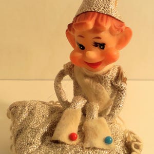 May include: A small elf figurine with a peach-colored face, wearing a silver textured hat and matching outfit. The elf sits on a silver quilted cushion with a white scalloped trim. The elf's white felt shoes feature one small red bead and one blue bead.