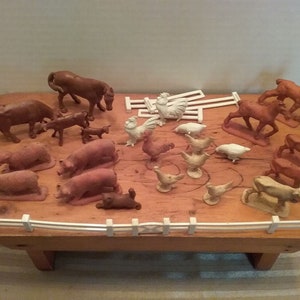 May include: A collection of miniature farm animal figurines, including cows, horses, pigs, chickens, and sheep, arranged on a wooden base with a white fence. The animals are primarily brown and tan.
