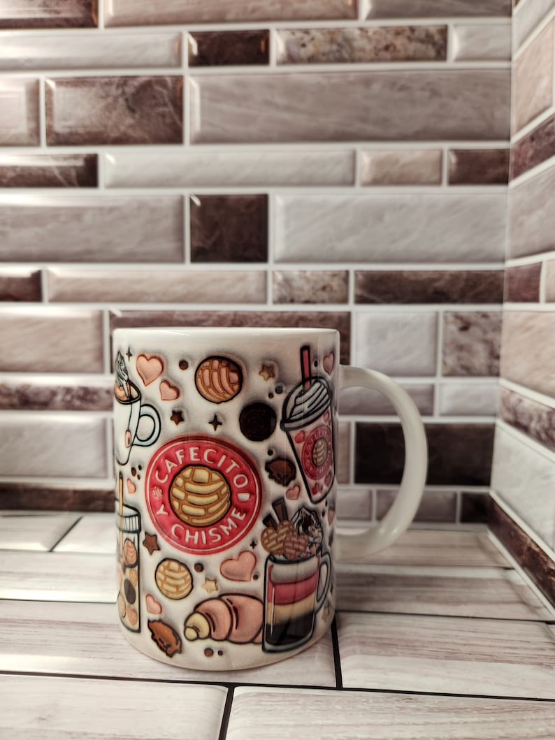 K&ouml;nnte beinhalten: Wei&szlig;e Keramiktasse mit einem farbenfrohen Design, das verschiedene mexikanische Geb&auml;cke, Getr&auml;nke und den Text "Cafecito y Chisme" zeigt.