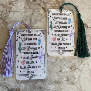 May include: Two white bookmarks with a green and purple tassel. The bookmarks feature a white background with black text and illustrations of herbs and flowers. The text reads "Always throw spilled salt over your left shoulder. Keep Rosemary by your garden gate. Plant Lavender for luck. Fall in love whenever you can."