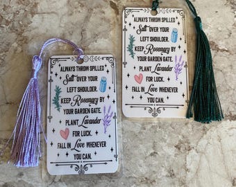 May include: Two white bookmarks with a green and purple tassel. The bookmarks feature a white background with black text and illustrations of herbs and flowers. The text reads "Always throw spilled salt over your left shoulder. Keep Rosemary by your garden gate. Plant Lavender for luck. Fall in love whenever you can."