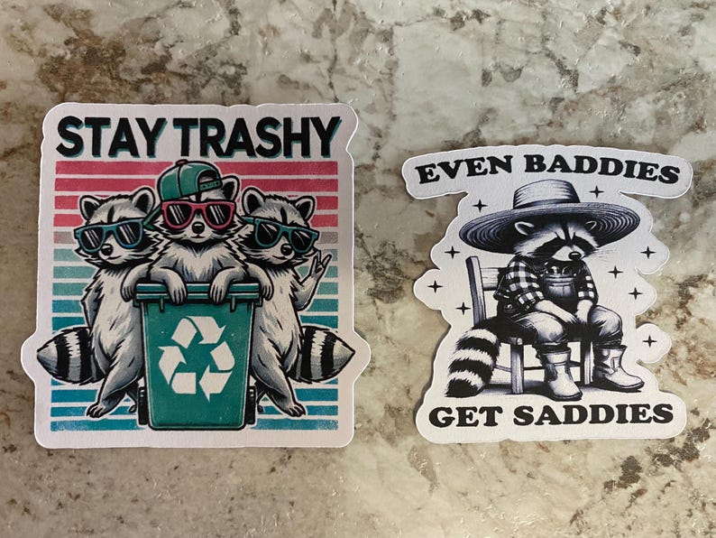 Puede incluir: Dos pegatinas con ilustraciones de mapaches. Una muestra tres mapaches con gafas de sol y el texto "STAY TRASHY". La otra representa un mapache con sombrero y peto con el texto "EVEN BADDIES GET SADDIES".