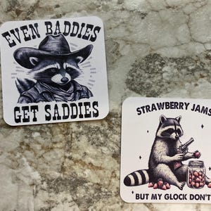 Puede incluir: Dos pegatinas cuadradas con ilustraciones de mapaches. Una presenta un mapache con sombrero de vaquero con el texto "EVEN BADDIES GET SADDIES". La otra muestra un mapache con una pistola y un tarro de fresas, con el texto "STRAWBERRY JAMS BUT MY GLOCK DON'T."