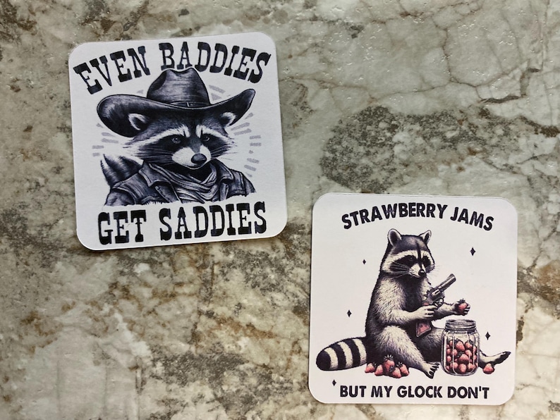 Puede incluir: Dos pegatinas cuadradas con ilustraciones de mapaches. Una presenta un mapache con sombrero de vaquero con el texto "EVEN BADDIES GET SADDIES". La otra muestra un mapache con una pistola y un tarro de fresas, con el texto "STRAWBERRY JAMS BUT MY GLOCK DON'T."