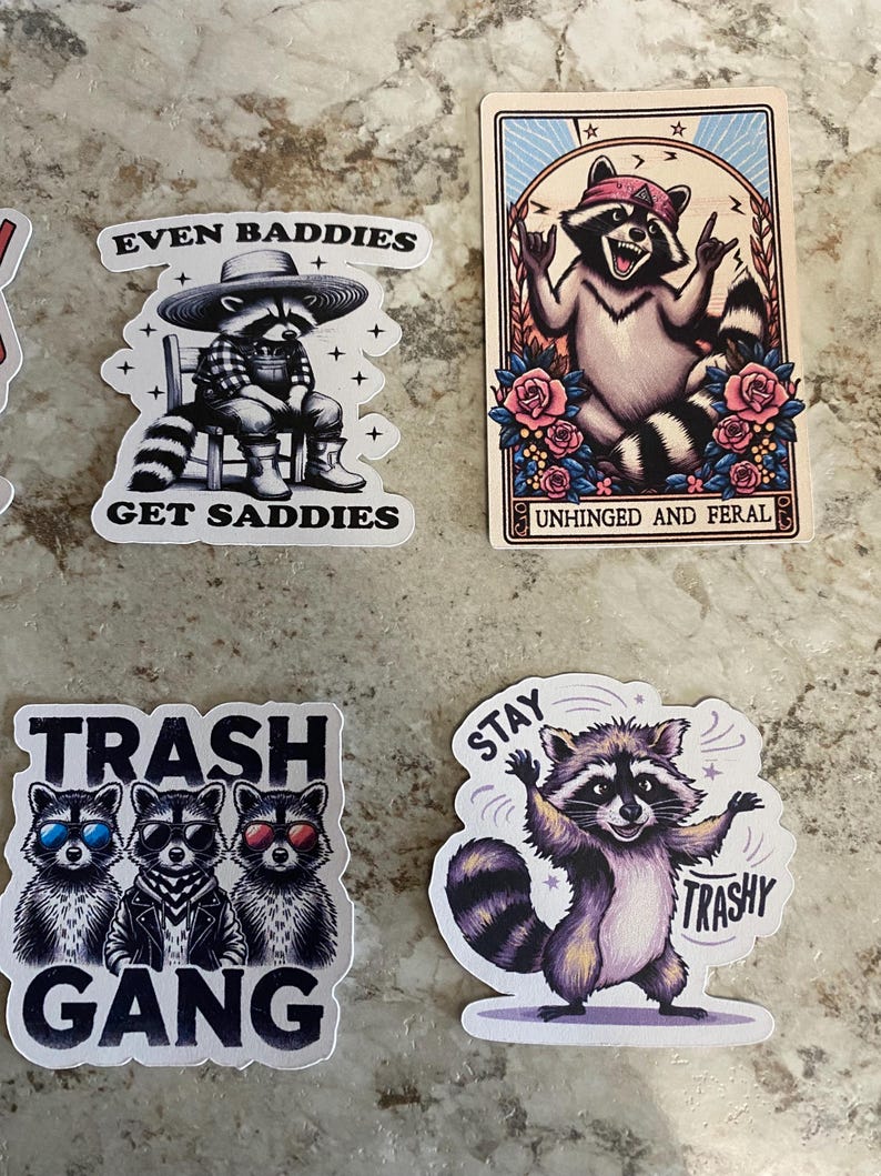 Puede incluir: Una colecci&oacute;n de pegatinas con tem&aacute;tica de mapaches. Una pegatina presenta un mapache con sombrero y botas con el texto "EVEN BADDIES GET SADDIES". Otra muestra una carta del tarot de mapache con el texto "UNHINGED AND FERAL". Una tercera pegatina dice "TRASH GANG", y la &uacute;ltima dice "STAY TRASHY".