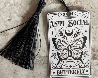 Può includere: Un segnalibro bianco con un'illustrazione in bianco e nero di una farfalla. Il segnalibro ha un fiocco nero e il testo "Anti-Social Butterfly" stampato su di esso.