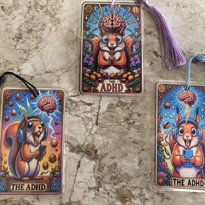 May include: Three colorful bookmarks featuring cartoon squirrels with the text "THE ADHD". Each bookmark has a different design, including a squirrel with a brain, headphones, and holding a phone. The bookmarks have tassels.
