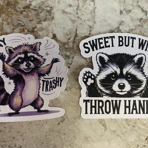 Puede incluir: Dos pegatinas con ilustraciones de mapaches. Una muestra un mapache morado con el texto "Stay Trashy". La otra pegatina representa un mapache con el texto "Sweet But Will Throw Hands."