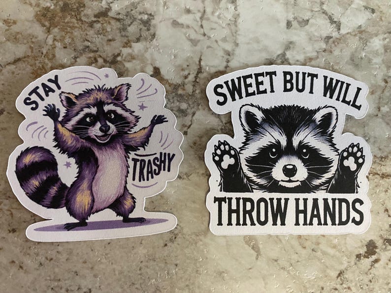 Puede incluir: Dos pegatinas con ilustraciones de mapaches. Una muestra un mapache morado con el texto "Stay Trashy". La otra pegatina representa un mapache con el texto "Sweet But Will Throw Hands."