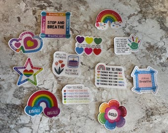 Puede incluir: Un conjunto de 11 parches de fieltro coloridos con diseños de arcoíris y texto relacionado con ejercicios de respiración. Los parches incluyen frases como "Stop and Breathe", "Try to Find Things You Can See", "Smell the Flowers", y "Hold".