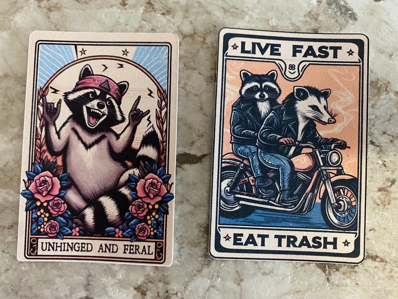 Puede incluir: Dos pegatinas rectangulares con dise&ntilde;os de mapaches y zarig&uuml;eyas de dibujos animados. Una presenta un mapache con un pa&ntilde;uelo rosa, el texto "UNHINGED AND FERAL" y rosas. La otra muestra un mapache y una zarig&uuml;eya en una motocicleta con el texto "LIVE FAST EAT TRASH".