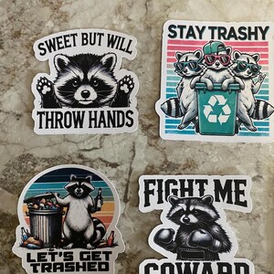 Puede incluir: Cuatro pegatinas con ilustraciones de mapaches y textos humor&iacute;sticos. Una dice "Sweet But Will Throw Hands". Otra dice "Stay Trashy" con mapaches con gafas de sol. Una tercera dice "Let's Get Trashed". La &uacute;ltima dice "Fight Me Coward."