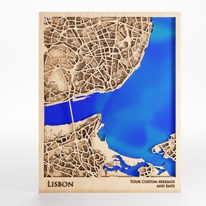 May include: Wooden laser-cut map of Lisbon, Portugal, with a blue river and surrounding city details. The map is framed in a light wood, with the city name and space for a custom message and date at the bottom.