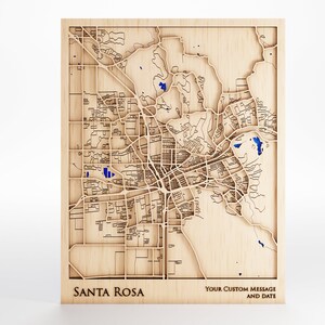 May include: A wooden map of Santa Rosa, California, with blue accents. The map is laser cut and features the city's streets and landmarks. The text "YOUR CUSTOM MESSAGE AND DATE" is printed below the map.