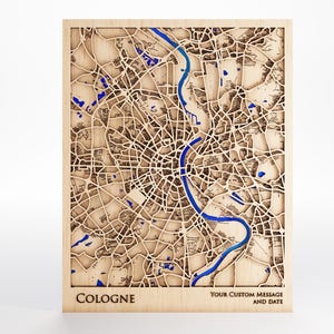 Puede incluir: Un mapa de madera de Colonia, Alemania, con detalles intrincados cortados con láser. El mapa presenta un río azul y detalles en azul que representan cuerpos de agua. La palabra "COLOGNE" está en la parte inferior, con espacio para un mensaje personalizado y una fecha.