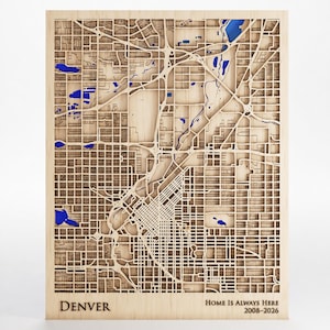May include: A rectangular wooden wall map of Denver featuring intricate laser-cut street grids and layered topography. The design includes vibrant blue accents for water features, with the text "DENVER" and "HOME IS ALWAYS HERE 2008-2026" printed.