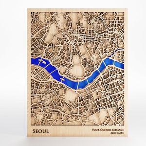 Può includere: Una mappa di legno di Seoul, Corea del Sud, con un fiume blu che la attraversa. La mappa è tagliata al laser e ha un aspetto rustico e naturale. Il testo "SEOUL" è stampato in basso a sinistra, e "YOUR CUSTOM MESSAGE AND DATE" è stampato in basso a destra.