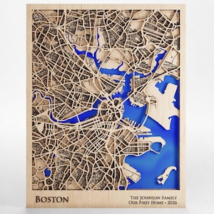 May include: A rectangular wooden wall map of Boston featuring intricate laser-cut street patterns in light wood tones layered over a vibrant blue water base. The text BOSTON is at the bottom left, and THE JOHNSON FAMILY OUR FIRST HOME 2026 is at the bottom right.