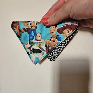 May include: A triangular fabric patch with a blue background and a colourful image of characters from the Toy Story movie franchise. The patch is sewn to a black and white polka dot fabric.