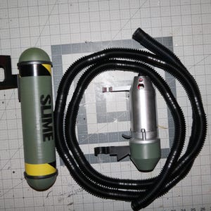 Könnte beinhalten: Ein grüner und gelber Schleimtank mit einem schwarzen Schlauch, der an einem silbernen und grünen Schleimwerfer befestigt ist. Der Tank trägt die Aufschrift "SLIME".