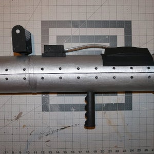 May include: A silver-colored prop weapon with a black handle and various black attachments. The cylindrical body has small black rivets and a nozzle with multiple holes. A black sight is mounted on top.