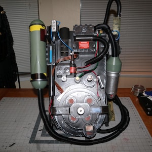 Könnte beinhalten: Ein silberner und schwarzer Protonenrucksack mit einem grün-schwarzen Schlauch, einem schwarzen Schlauch und einem silbernen und schwarzen runden Gerät. Der Rucksack steht auf einer Holzoberfläche.