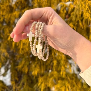 May include: Three stacked bracelets with white cross charms and beige beads.