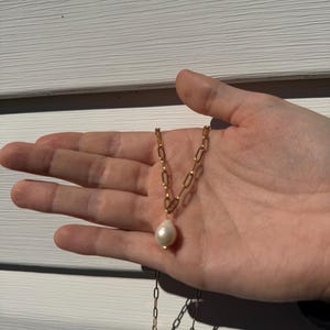 May include: A gold-colored chain necklace with a large, teardrop-shaped white pearl pendant. The necklace is displayed on a hand, showcasing the delicate chain links and the luster of the pearl.