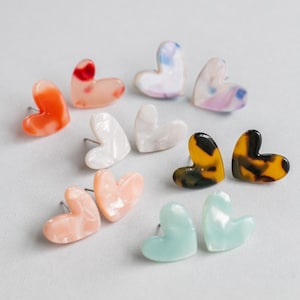 May include: A collection of heart-shaped stud earrings in various colors and patterns. The earrings include shades of orange, pink, white, blue, and tortoiseshell. The earrings are displayed on a white surface.