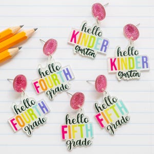 May include: Clear acrylic earrings with pink glitter studs. The earrings feature the phrases "hello KINDERGARTEN", "hello FOURTH grade", and "hello FIFTH grade" in colorful block letters. Pencils are in the background.