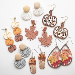 May include: A collection of fall-themed earrings, including pumpkin, maple leaf, and coffee cup designs. The earrings feature various colors, textures, and patterns, such as leopard print and woven details. The earrings are displayed on a white surface.