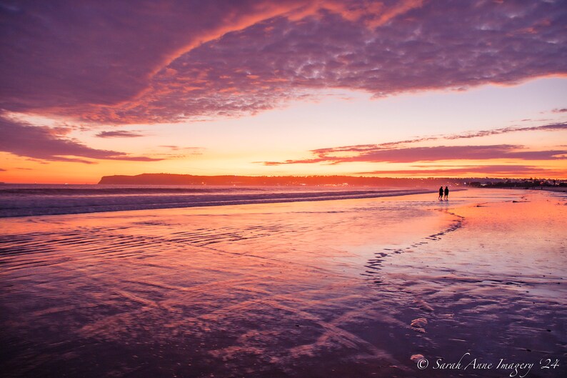 May include: A couple walks along a sandy beach at sunset. The sky is a vibrant mix of pink, orange, and purple, reflecting in the wet sand.