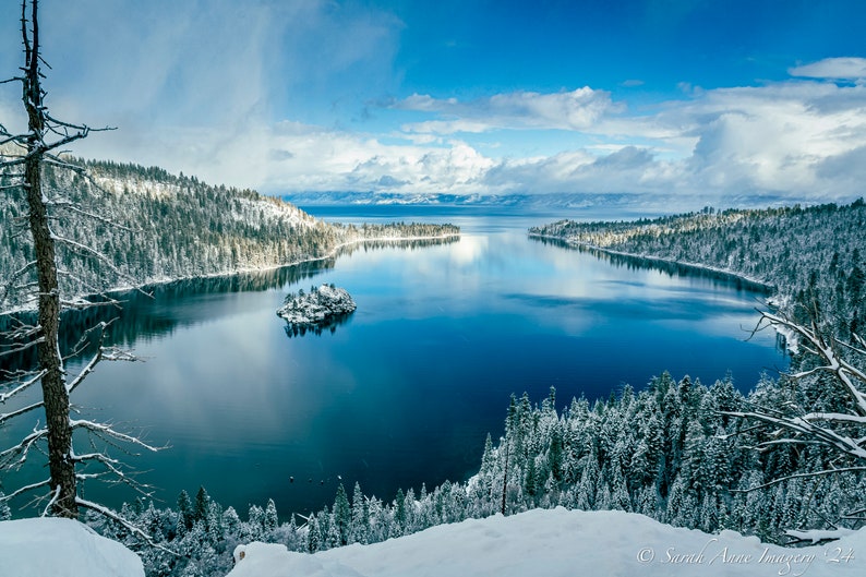 Puede incluir: Una vista panor&aacute;mica de un lago de monta&ntilde;a cubierto de nieve con una peque&ntilde;a isla en el centro. El agua es de un azul profundo y el cielo es de un azul brillante con nubes blancas. Los &aacute;rboles en la orilla est&aacute;n cubiertos de nieve.