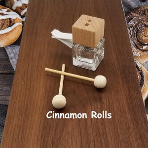 May include: A wooden car air freshener with a glass bottle and two wooden sticks. The air freshener is on a wooden surface with the text "Cinnamon Rolls" written below. There are also cinnamon rolls on a brown linen cloth.