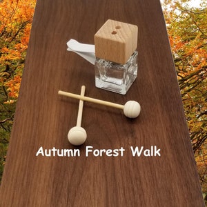 以下が含まれることがあります： 木製の蓋と白いボールの付いた木製の棒が2本付いたガラス瓶。瓶には「Autumn Forest Walk」とラベルが貼られています。背景はぼやけた秋の森の画像です。
