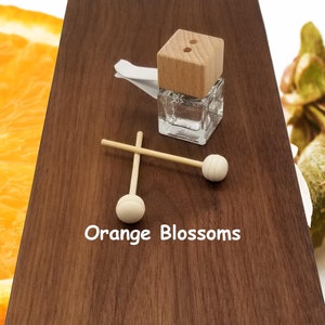 May include: A small glass bottle with a wooden lid and two wooden sticks with round ends. The bottle is filled with a clear liquid. The text "Orange Blossoms" is printed on a wooden surface.