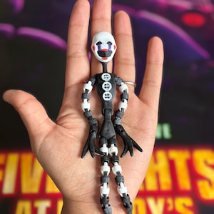 Może przedstawiać: 3D-drukowana, przegubowa figurka Puppet, z białą głową, czarnymi detalami twarzy, czarnym korpusem i czarno-białymi kończynami. Figurka ma trzy białe guziki na piersi i jest trzymana w dłoni. Tło zawiera tekst "Five Nights at Freddy's".