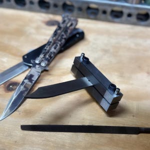 Op de afbeelding: Een zwarte metalen messen-slijpbeugel met een vastgeklemd mes. Een zwart en camouflage vouwmes en een zwarte vijl zijn ook op de afbeelding te zien.