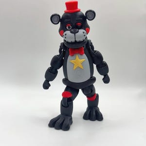 May include: A black and gray articulated bear figure with a red top hat, bow tie, and knee pads. The bear has a gray chest with a gold star and round ears. The figure is standing on a white surface.