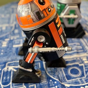 May include: A robotic figure featuring a black and orange cylindrical body with a clear dome top, black circular accents, and a silver-colored arm attachment extending from the side, positioned next to a secondary green and silver robotic figure.